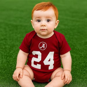 May include: A baby in a maroon bodysuit with the number 24 in white and a white letter "A" inside a circle. The baby has red hair and is sitting on green grass.