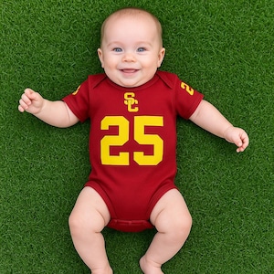 Baby lying on grass wearing a maroon football-jersey-style onesie with gold details, featuring the USC Trojans logo and number 25 on the front.