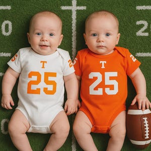 Two babies lying on a football field backdrop. One wears a white football-style onesie with orange details and the number 25 below the Tennessee Vols logo, while the other wears an orange version of the same onesie. A small football rests beside the baby in orange.