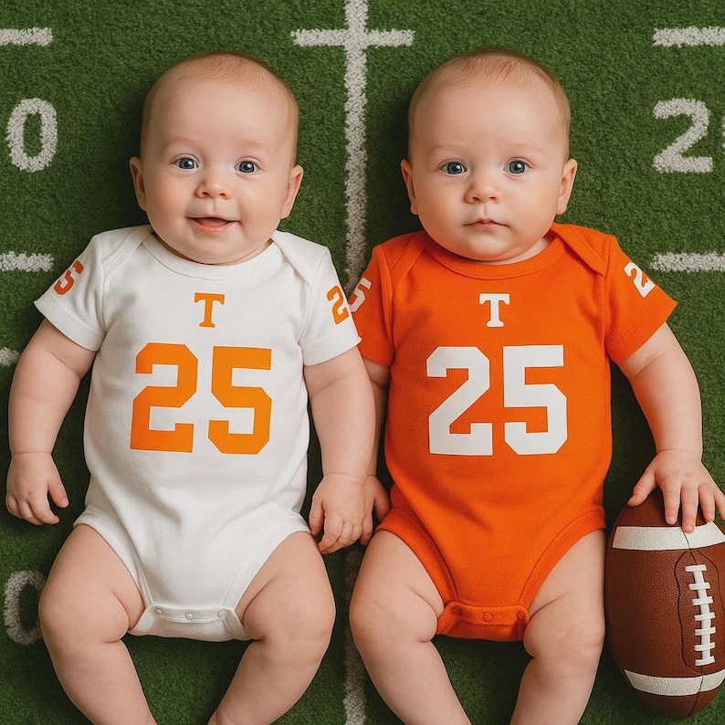 Two babies lying on a football field backdrop. One wears a white football-style onesie with orange details and the number 25 below the Tennessee Vols logo, while the other wears an orange version of the same onesie. A small football rests beside the baby in orange.