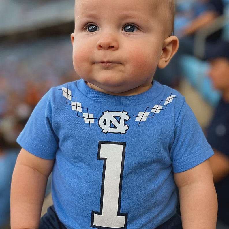 A baby wearing a light blue t-shirt with a white and blue diamond pattern on the collar. The shirt has a large white number 1 on the front and a University of North Carolina logo in the center. The baby has fair skin and blue eyes.