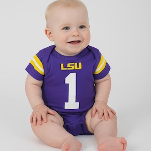 Smiling baby sitting on a plain white backdrop, wearing a purple football-style onesie with yellow striping on the sleeves. The front of the onesie features the LSU logo above a large white number 1.