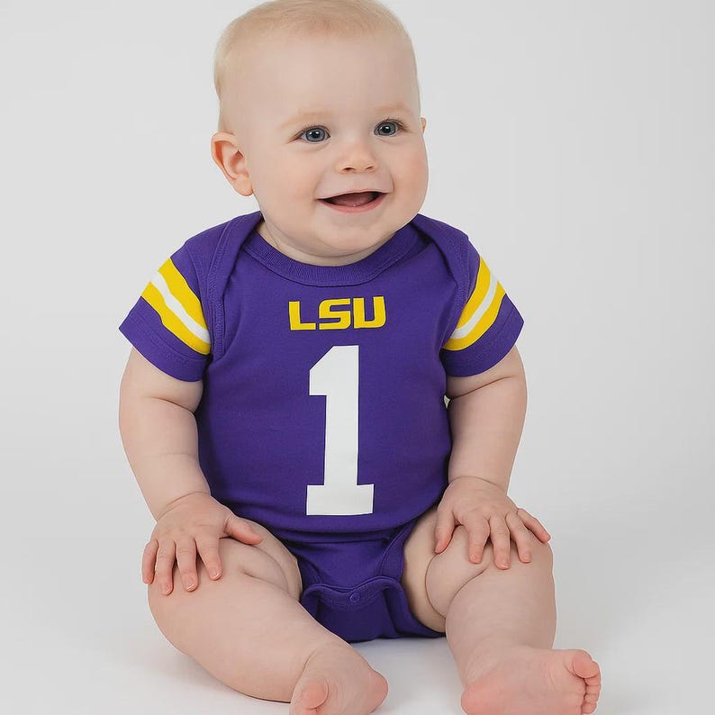 Smiling baby sitting on a plain white backdrop, wearing a purple football-style onesie with yellow striping on the sleeves. The front of the onesie features the LSU logo above a large white number 1.