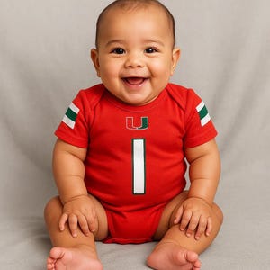 May include: A smiling baby wearing a red onesie with a white number 1 and green and white stripes on the sleeves. The onesie has a green and orange U logo on the chest.