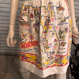 May include: A white apron featuring a colorful map of Florida with illustrations of landmarks and the text "FLORIDA The Sunshine State." The apron has a yellow and red border and a white waistband.