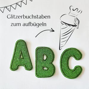 Könnte beinhalten: Drei grüne Glitzer-Aufbügler in Form der Buchstaben A, B und C. Das Bild enthält eine schwarze Strichzeichnung einer Eistüte sowie den Text Glitzerbuchstaben zum aufbügeln mit einem geschwungenen Pfeil, der auf die Eistüte zeigt.