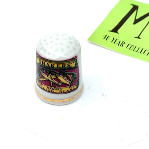 Può includere: Ditale in ceramica bianca con bordo dorato e un'immagine colorata di una lattina di fagiolini in scatola Shaker's.