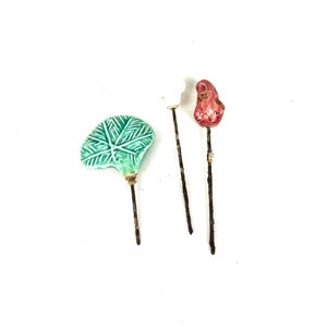 May include: Three ceramic cake toppers, one shaped like a leaf in green glaze, one shaped like a flower in red glaze, and one shaped like a white flower. The toppers are attached to thin metal wires.