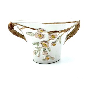 May include: White ceramic planter with embossed floral design. The planter features a detailed relief of pink flowers and green leaves. It has two brown handles and a decorative rim. The planter is ideal for small plants or flowers.