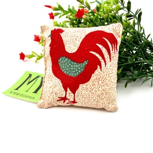 May include: A small, square, cream-coloured pillow with a red rooster design. The rooster has a green chest and a white background with small red flowers.