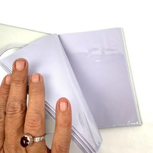 Peut inclure: Un album photo blanc avec des pochettes en plastique transparent. L'album est ouvert, r&eacute;v&eacute;lant plusieurs pages de pochettes. L'album est tenu par une main portant une bague en argent avec une pierre rouge.