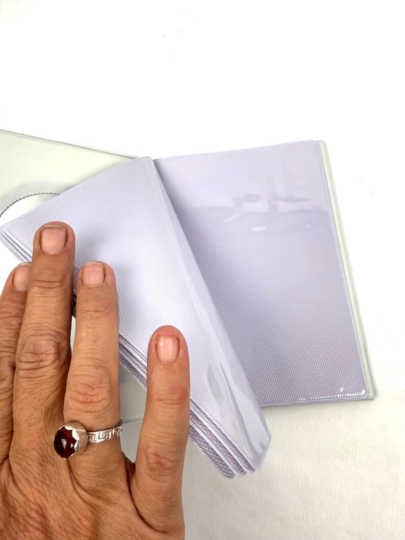 Peut inclure: Un album photo blanc avec des pochettes en plastique transparent. L'album est ouvert, r&eacute;v&eacute;lant plusieurs pages de pochettes. L'album est tenu par une main portant une bague en argent avec une pierre rouge.