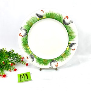 May include: White ceramic dinner plate with a green and white border featuring a repeating pattern of chickens and grass. The plate has a raised rim and a smooth surface.