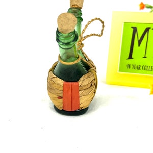 May include: Two small green glass bottles with cork tops are tied together with twine and wrapped in a woven straw basket with a red stripe.