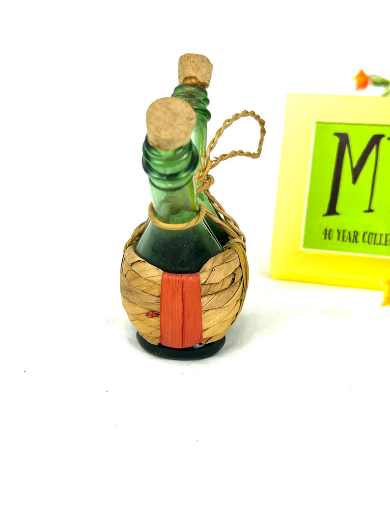 May include: Two small green glass bottles with cork tops are tied together with twine and wrapped in a woven straw basket with a red stripe.