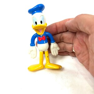Może przedstawiać: Plastikowa figurka Donalda Ducka w ż&oacute;łtym i białym kolorze, z niebieskim kapeluszem marynarskim i czerwoną muszką. Figurka stoi z wyciągniętymi rękami.