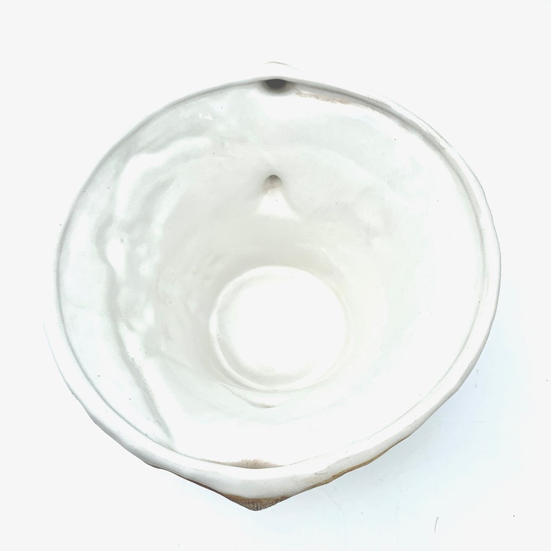 May include: A white ceramic bowl with a rounded shape and a small spout. The bowl has a smooth, glossy interior and a slightly textured exterior. The bowl is empty and appears to be clean.