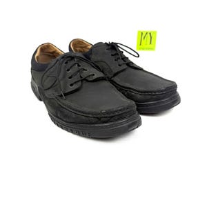 May include: A pair of black leather shoes with laces. The shoes have a rounded toe and a thick sole. The interior of the shoe is a light brown color. A yellow sticker with the letters "MY" is in the background.