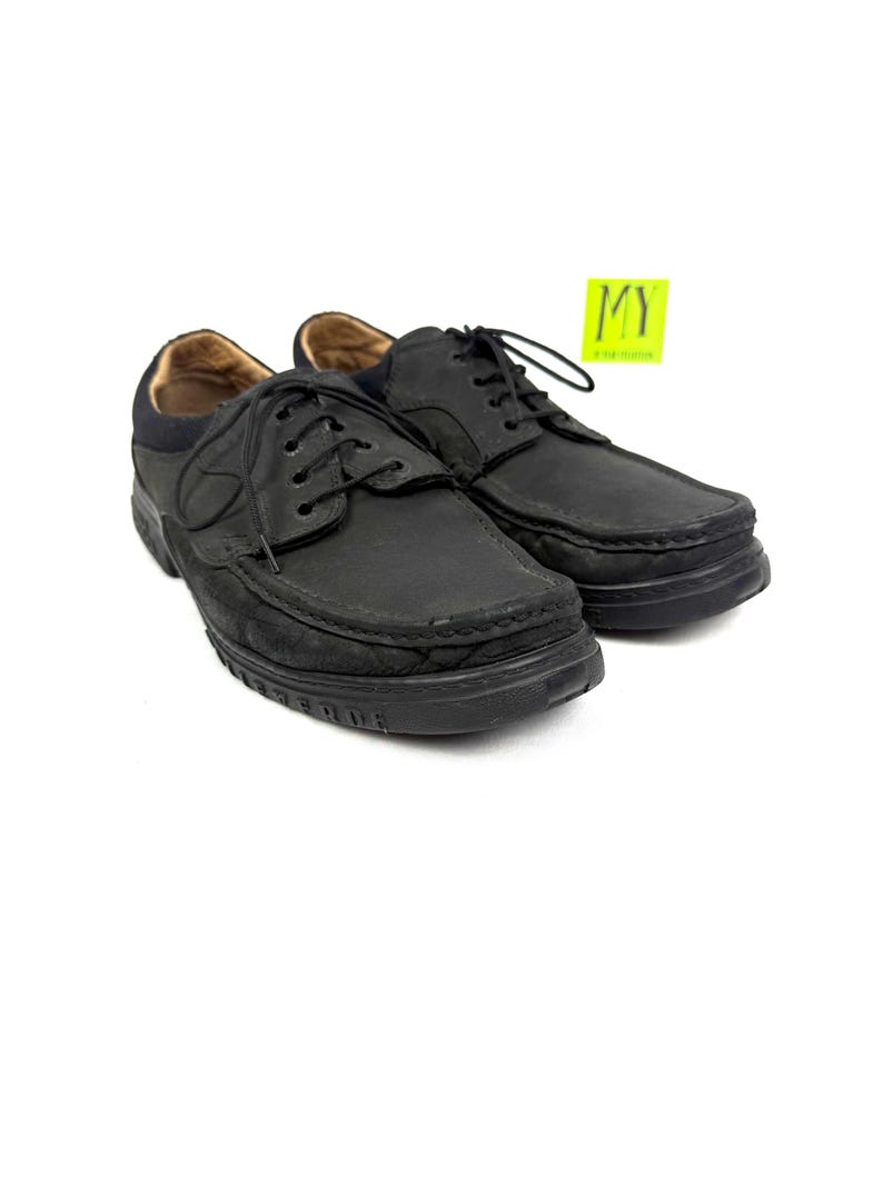 May include: A pair of black leather shoes with laces. The shoes have a rounded toe and a thick sole. The interior of the shoe is a light brown color. A yellow sticker with the letters "MY" is in the background.