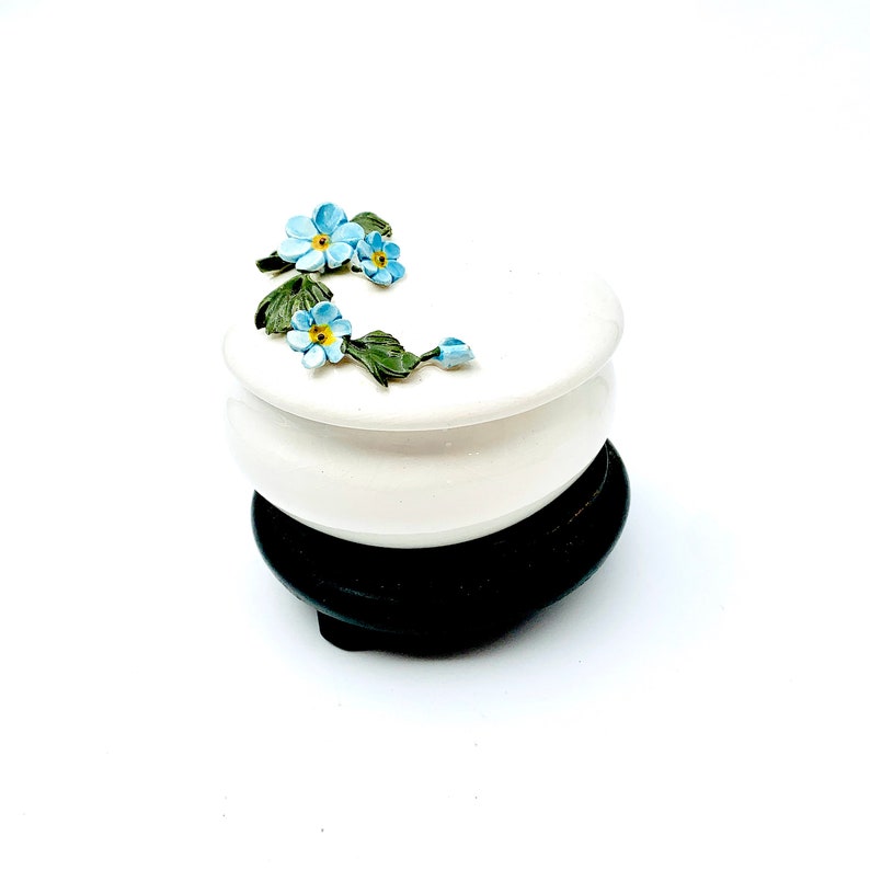 May include: White ceramic trinket box with a floral design featuring blue forget-me-not flowers and green leaves. The box sits on a black wooden base.