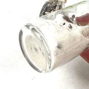 May include: Two clear glass bottles filled with white sand and seashells. The bottles are on a white surface.