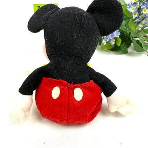 May include: A plush Mickey Mouse toy, viewed from the back, features a black head and ears, red shorts with white buttons, and white gloves. The toy has a yellow shirt and is set against a white background.