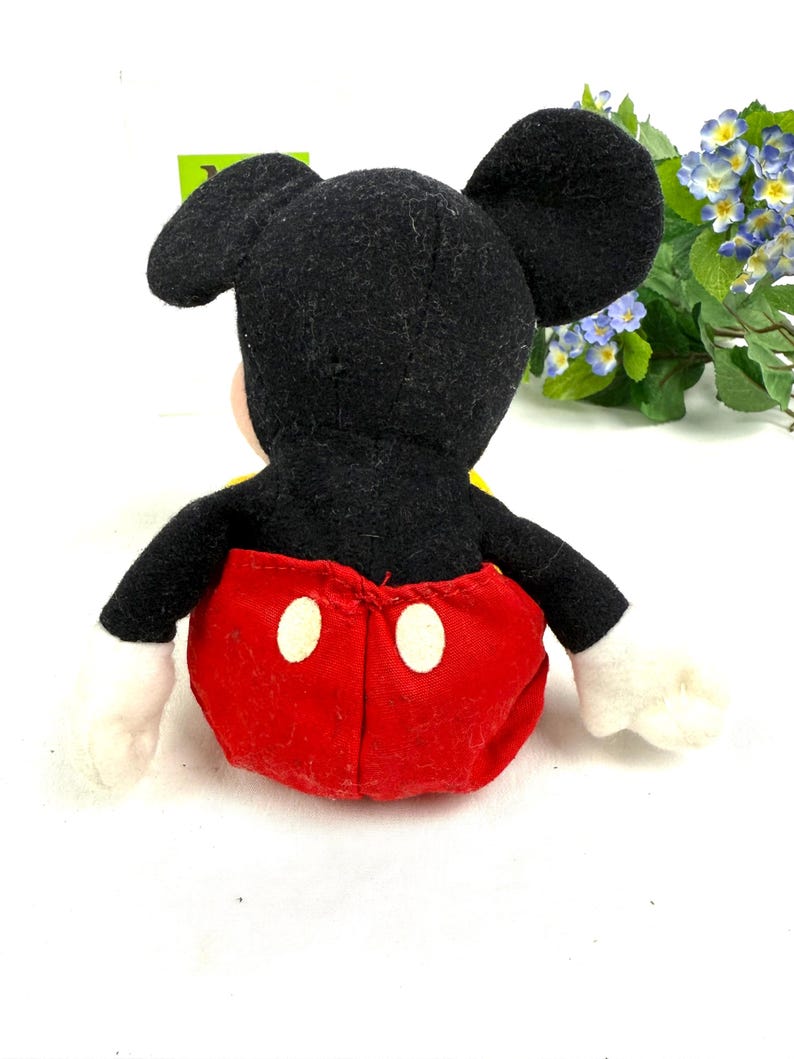 May include: A plush Mickey Mouse toy, viewed from the back, features a black head and ears, red shorts with white buttons, and white gloves. The toy has a yellow shirt and is set against a white background.