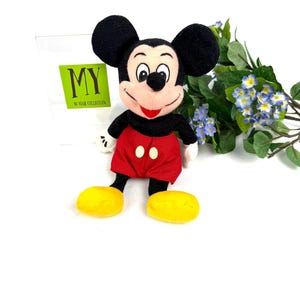 May include: A plush Mickey Mouse toy with black ears, a red shorts, yellow shoes, and white gloves. The toy is smiling and has a red mouth. The background includes a sign that says "MY 46 YEAR COLLECTION" and some blue flowers.
