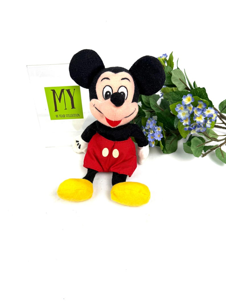 May include: A plush Mickey Mouse toy with black ears, a red shorts, yellow shoes, and white gloves. The toy is smiling and has a red mouth. The background includes a sign that says "MY 46 YEAR COLLECTION" and some blue flowers.
