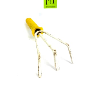 Pode incluir: Um pequeno ancinho de mão de jardim com três dentes. Os dentes são brancos e ligeiramente enferrujados. O cabo é amarelo com uma pega texturizada. Um autocolante verde com o texto "MY 90 YEAR COLLECTION" está no fundo.