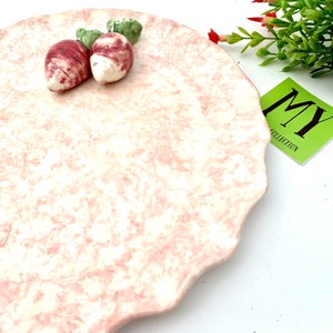 May include: A pink ceramic serving plate with a scalloped edge. The plate has a marbled design and features two small, red and green ceramic radishes.