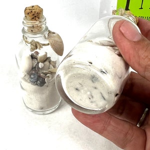 May include: Two small glass bottles filled with white sand and seashells. The bottles have cork stoppers and are tied with twine.