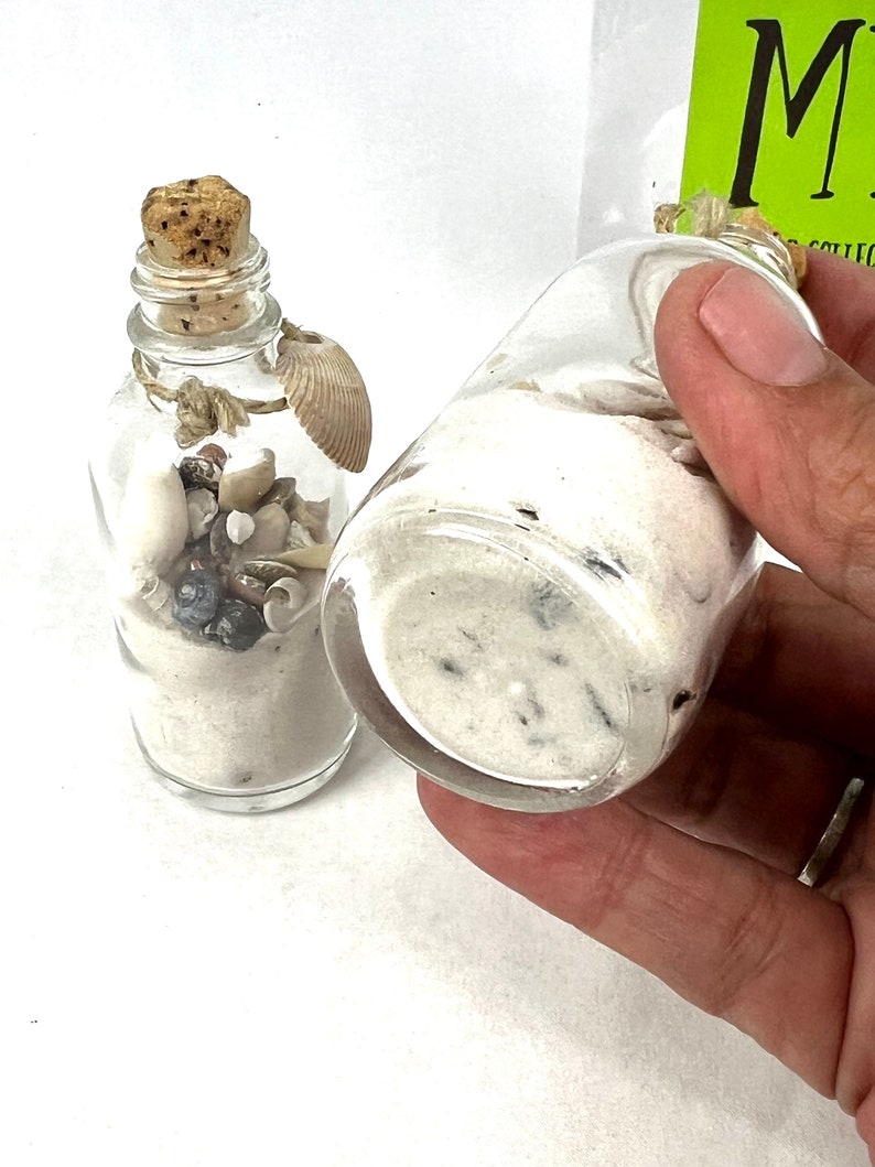 May include: Two small glass bottles filled with white sand and seashells. The bottles have cork stoppers and are tied with twine.