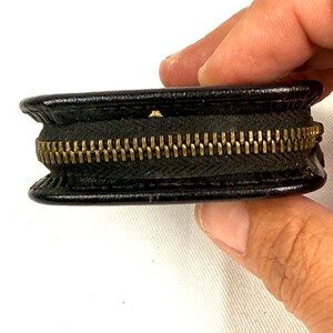 May include: A small, black leather coin purse with a brass zipper. The purse is round and has a classic design. The zipper is a prominent feature, adding a touch of vintage style. The purse is held by a person's hand.