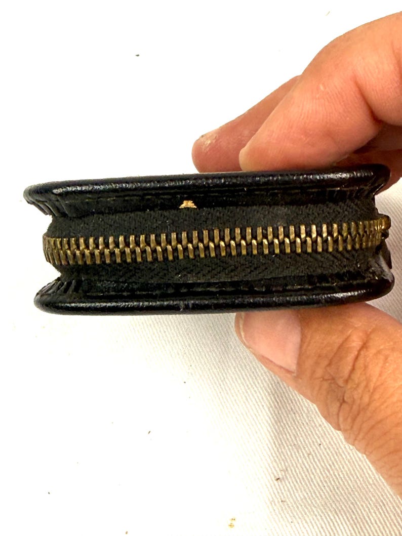 May include: A small, black leather coin purse with a brass zipper. The purse is round and has a classic design. The zipper is a prominent feature, adding a touch of vintage style. The purse is held by a person's hand.