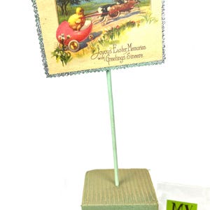 May include: An Easter-themed display featuring a vintage postcard with a chick in a red carriage pulled by rabbits. The postcard has a glittery border and is mounted on a green stick and a green foam base. Text reads "Joyous Easter Memories with Greetings Sincere."