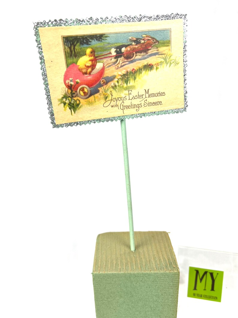 May include: An Easter-themed display featuring a vintage postcard with a chick in a red carriage pulled by rabbits. The postcard has a glittery border and is mounted on a green stick and a green foam base. Text reads "Joyous Easter Memories with Greetings Sincere."