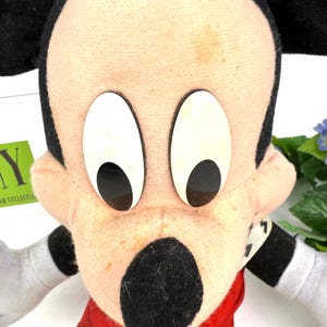 May include: A vintage Mickey Mouse plush toy. The plush features a black nose, ears, and arms, with a red shirt and white gloves. The face is beige with large white eyes and black pupils. The toy is a classic Disney character.