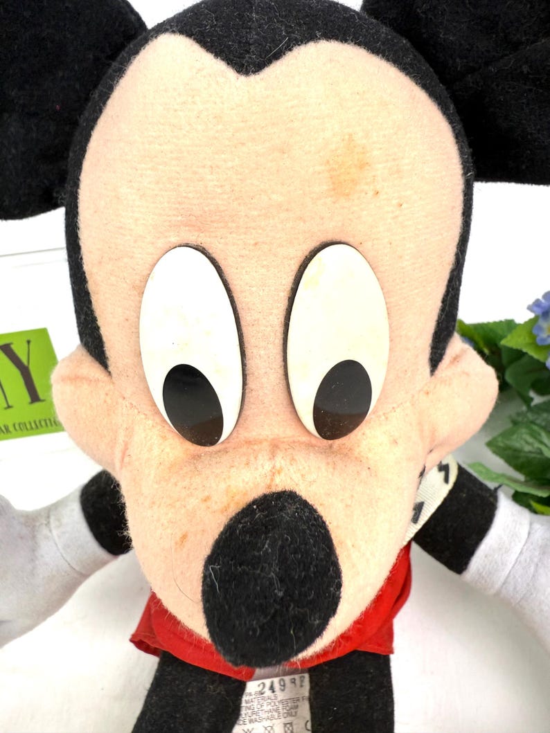May include: A vintage Mickey Mouse plush toy. The plush features a black nose, ears, and arms, with a red shirt and white gloves. The face is beige with large white eyes and black pupils. The toy is a classic Disney character.