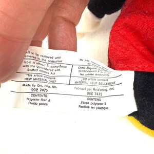 May include: A close-up of a white tag with black text, held by a hand. The tag reads "NEW MATERIAL ONLY" and lists contents as polyester fiber and plastic pellets. The tag is affixed to a red and black fabric.