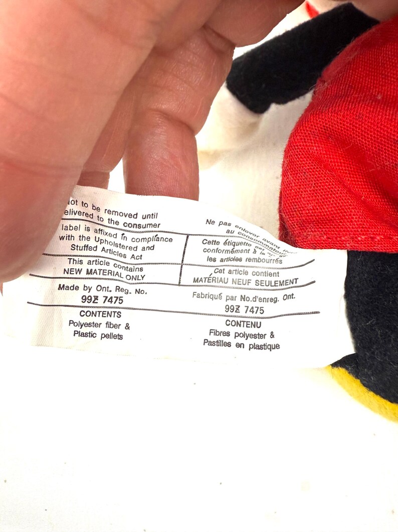 May include: A close-up of a white tag with black text, held by a hand. The tag reads "NEW MATERIAL ONLY" and lists contents as polyester fiber and plastic pellets. The tag is affixed to a red and black fabric.