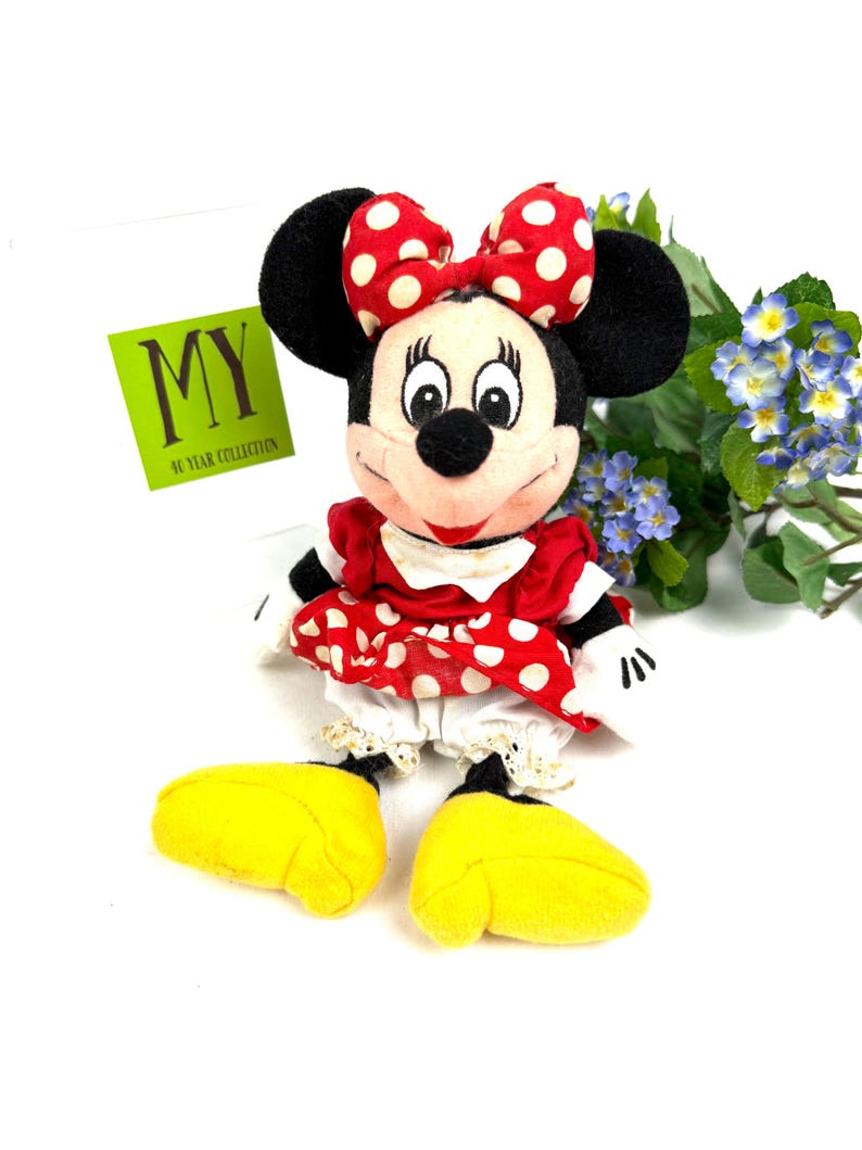 May include: A Minnie Mouse plush doll with a red and white polka dot bow, dress, and yellow shoes. The doll has black ears and a smiling face. The tag reads "MY 40 YEAR COLLECTION".