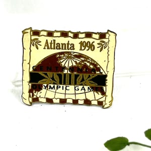 Può includere: Uno spillo dorato con sfondo bianco e un globo rosso al centro. Lo spillo presenta il testo "Atlanta 1996" e "Centennial Olympic Games".