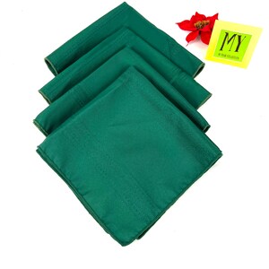 May include: Set of four green linen napkins. The napkins are folded and stacked on a white background. The napkins have a subtle texture and a slightly darker green border.