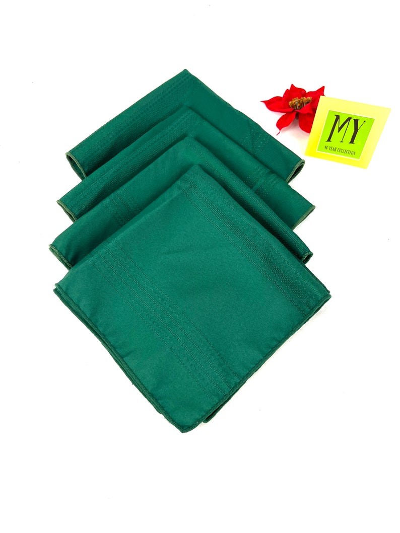 May include: Set of four green linen napkins. The napkins are folded and stacked on a white background. The napkins have a subtle texture and a slightly darker green border.