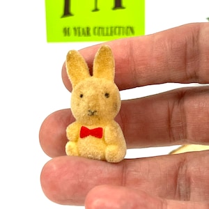 May include: A small, plush, tan bunny rabbit wearing a red bow tie. The bunny is sitting on a hand.