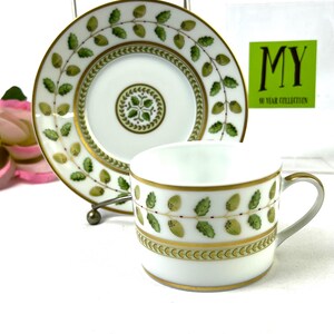 May include: A white porcelain teacup and saucer with a green and gold leaf design. The saucer has a gold rim and the cup has a gold band with a green and gold leaf design. The text "MY 40 YEAR COLLECTION" is visible on a white card in the background.