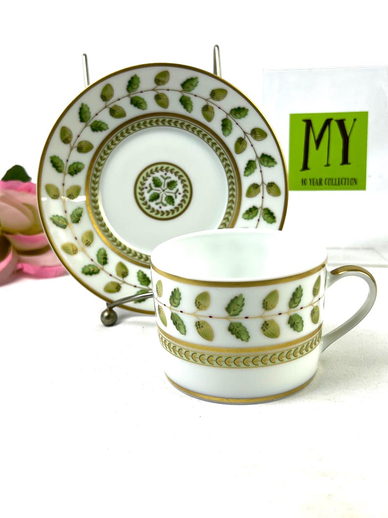 May include: A white porcelain teacup and saucer with a green and gold leaf design. The saucer has a gold rim and the cup has a gold band with a green and gold leaf design. The text "MY 40 YEAR COLLECTION" is visible on a white card in the background.