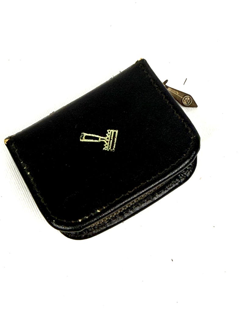 May include: Black leather coin purse with a gold-colored gavel emblem. The purse is small and rectangular with a rounded flap. A small metal tag is attached to the side. The purse is likely vintage.