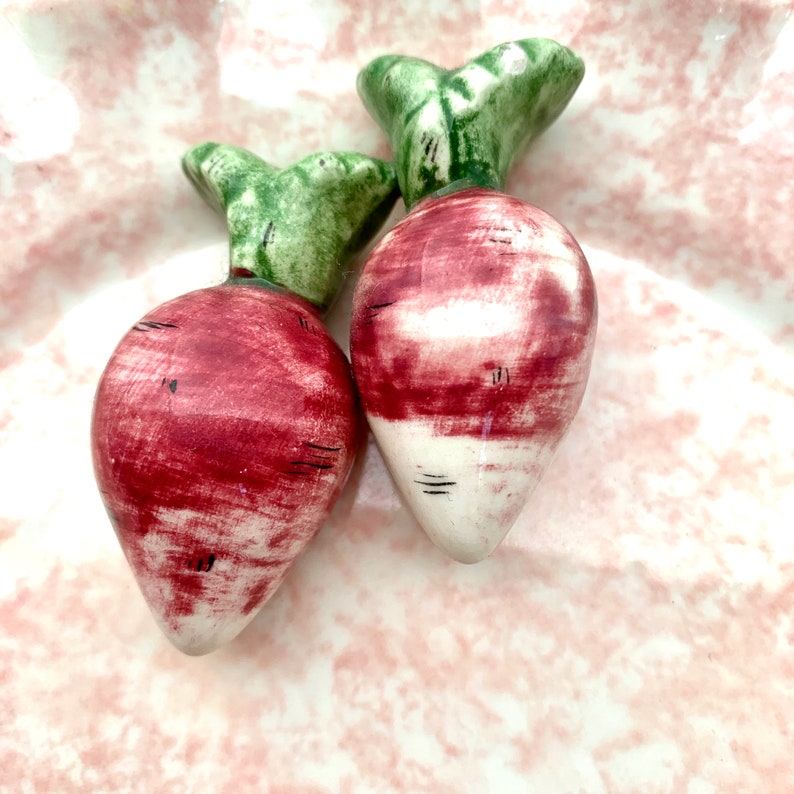 May include: Two ceramic radish figurines with green tops and red and white painted bodies. The radishes are sitting on a pink and white patterned surface.